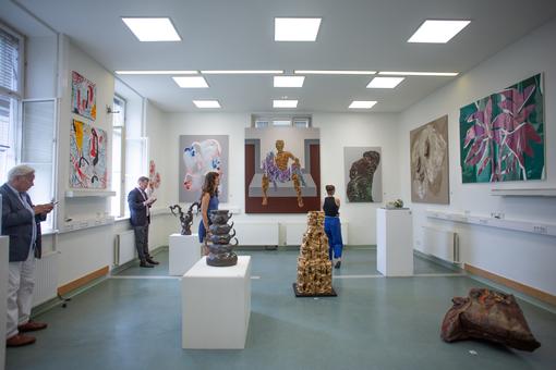 Ausstellungsansicht: unterschiedliche Gemälde an weißen Wänden, Skulpturen auf weißen Sockeln, dazwischen Besucher:innen