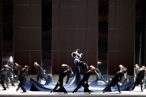 Szenefoto aus dem Ballett "Die Fledermaus" der Wiener Staatsoper. Tanzszene mit Tänzern in schwarzen Fracks, die eine Dame mit ausladendem Hut und Fächer umtanzen, indem sie die Enden ihres dunkelblauen extrem langen Kleides festhalten
