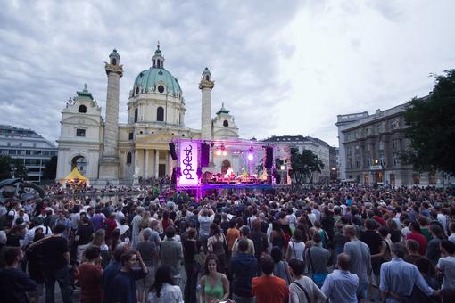 Foto der Bühne des Popfestes Wien, im Hintergrund die Karlskirche, vor der Bühne Publikum