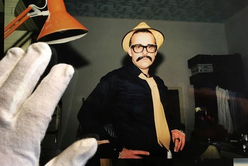 The press photo by Sabine Schwaighofer from the “Homo Diaries” series shows a man in dark clothing, with glasses, a beige hat and tie, looking into the camera, a hand in white gloves in the foreground