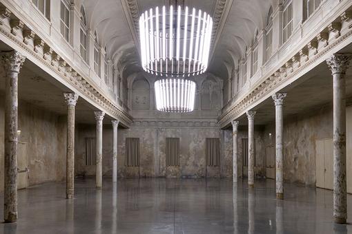 Foto zeigt einen historischen Saal mit korinthischen Säulen und Fenstern an der Seite, zwei moderne Luster mit Neonleuchten erhellen das historische Foyer