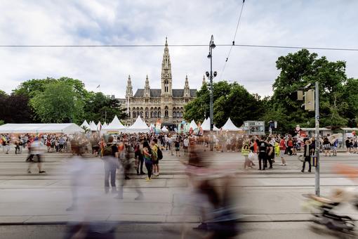 Das Foto zeigt das Wiener Rathaus vom Burgtheater aus gesehen, davor auf den Rathausplatz das Pride Village mit weißen Zelten und vielen Besucher:innen