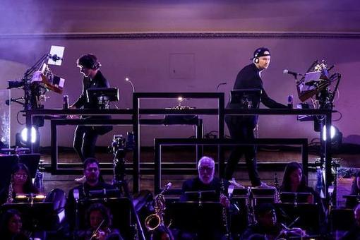 The photo shows the musicians Camo & Krooked with their music equipment on stage, in front of them some orchestra musicians, the stage is illuminated in the colors magenta and purple