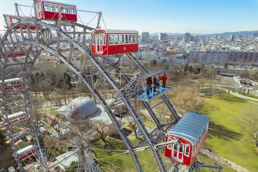 Foto des Wiener Riesenrad, Menschen auf einer Stahl-Glas-Plattform anstelle einer Gondel, im Hintergrund die Skyline von Wien