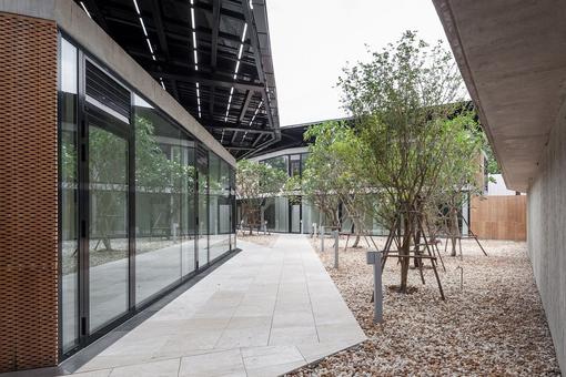 The photo shows the Austrian Embassy in Bangkok, a modern glass and steel construction with an inner courtyard planted with young trees