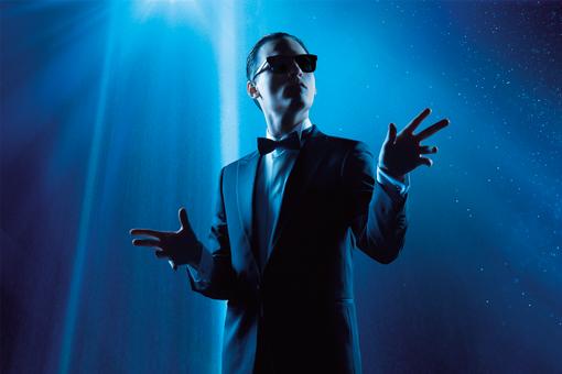 Subject photo showing the main actor of the musical in the style of the singer Falco in in a dark suit with bow tie and sunglasses, in the spotlight against a deep blue background