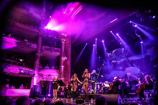 Foto mit Szene der Show auf der Bühne der Wiener Staatsoper, das Orchester im Hintergrund, drei Sängerinnen im Vordergrund, die Szene in magentalila Licht getaucht