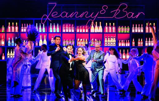 Szenefoto aus dem Musical, tanzende Protagonisten in einer Bar, im Mittelpunkt ein Paar in dunkler Abendgardarobe, das Richtung Kamera blickt