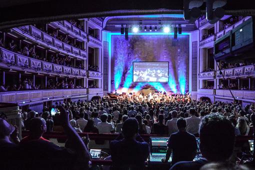 Foto mit Szene der Show auf der Bühne der Wiener Staatsoper, Blick vom violett beleuchteten Publikumsraum, vom Stehplatz Parterrre über das Publikum hinweg auf die Bühne