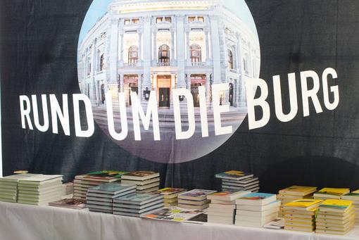 Logo der Veranstaltung: Der Schriftzug "Rund um die Burg" vor dem Bild des Burgtheaters mit schwarzem Hintergrund, im Vordergrund einige Stapel mit Büchern