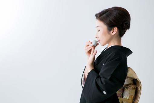 Foto einer Japanerin in einem taditionellen dunklen Kimono, die eine kleine japanische Tasse zum Mund führt 