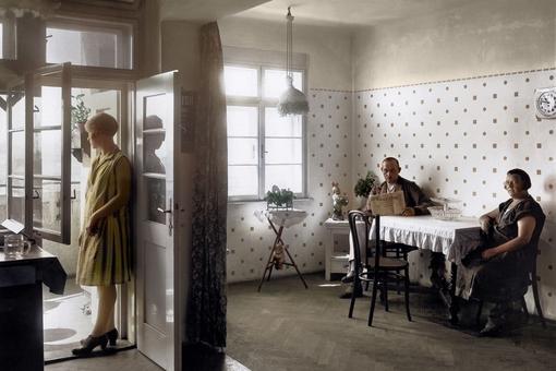 Photo of a modern working class apartment of 1920s, scene in a kitchen, a man reading newspaper and a woman sitting at a table with tablecloth, a second woman looks out on the balcony