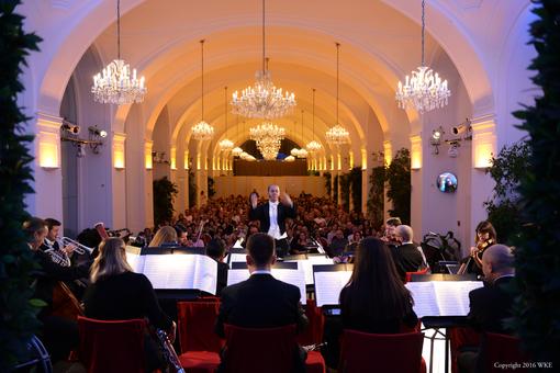 Das Foto zeigt das Schloss Schönbrunn Orchester von hinten, mit Blick auf den Dirigenten, im Hintergrund des Fotos die Orangerie des Schlosses Schönbrunn mit zahlreichen Kristalllustern und das Publikum
