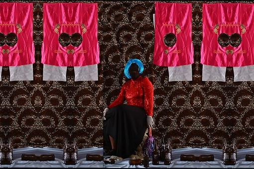 Eine bekleidete Puppe ohne Gesicht, die eine Frau darstellen soll, sitzt vor einem dunkel- und hellbraunen Wandteppich, vor dem pinke und weiiße Stoffstücke mit Löchern hängen