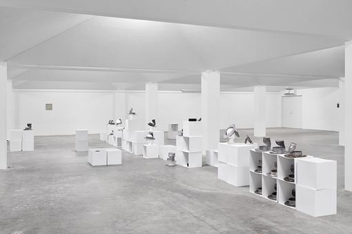 Exhibition view in black and white: cube-like white shelves and chests in which and on which different objects are placed