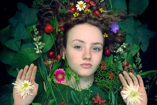 Photo of young woman whose head is framed by flowers and plants, in her palms white flower blossoms