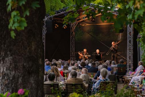The photo shows the open-air stage of the Summer Rhapsody in the garden of the Liechtenstein Garden Palace with three musicians playing a concert and the audience gathered in front of the stage