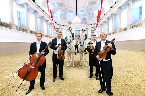 Photo in the Winter Riding School of the Spanish Riding School: two Lipizzaner horses with riders, in the foreground four members of the Vienna Philharmonic Orchestra in black tailcoats and instruments in hand