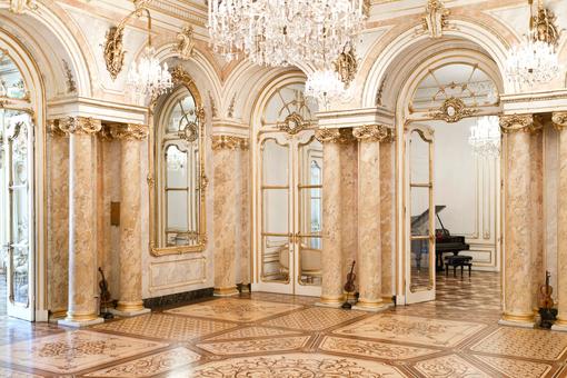 The photo shows the baroque Hall of Mirrors at Palais Coburg with its marble floor, marble columns, opulent chandeliers and gold-framed mirrors