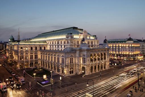 Zu sehen ist das historische Gebäude der Wiener Staatsoper in abendlicher Beleuchtung, von einem Gebäude gegenüber vom Ring aus fotografiert
