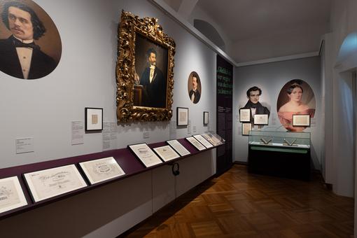 Das Foto zeigt eine Ausstellungsansicht, ein Raum mit Porträts der Familie Strauß an den Wänden, das Bild von Johann Strauss Sohn in einem prunkvollen goldenen Rahmen, darunter Vitrinen mit Originaldokumenten