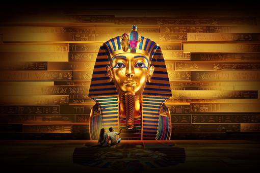 Bild der berühmten Totenmaske des Tutanchamun in übergroßer Darstellung, im Hintergrund eine Wand mit Hyroglyphen, vor der Maske sitzen eine Frau und ein Mann