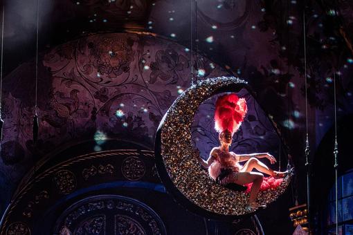 The scene photo from the opera “Tannhäuser” shows a glittering moon hanging above the stage. One of the opera singers is sitting on it, dressed like a revue dancer with a large red headdress. The scene is lit from behind.