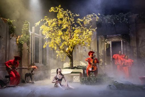 Scene photo from the production “The Grief of Red Granny” of the Vienna Festival 2025. The scene shows the inner courtyard of a building, with a bed from which a tree grows in the middle, surrounded by performers in red costumes