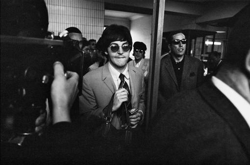 Black and white photo of Paul McCartney surrounded by a group of people, in the foreground a cameraman
