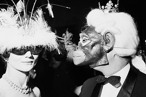 Schwarzweiß-Foto einer eleganten Dame mit Federhut und schwarzer Sonnenbrille, begleitet von einem Mann in schwarzem Anzug mit weißer Perücke und Krönchen mit einer Affenmaske im Gesicht