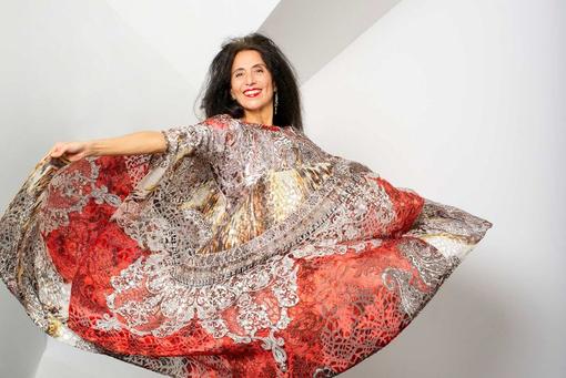 The photo shows the artist Timna Brauer in an orange-beige, slightly golden, oriental dress, which she is swinging around her body with her arms, wearing her long dark hair loose and smiling at the camera