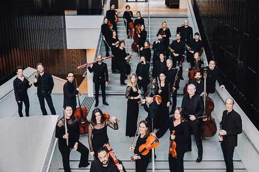 The photo shows the members of the Tyrolean Chamber Orchestra InnStrumenti dressed in black with their instruments in their hands standing scattered in a staircase of a modern building