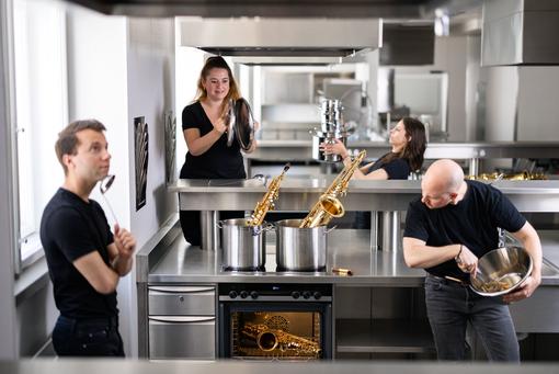Das Foto zeigt das Aureum Saxophon Quartett in einer Küche eines Gastronomiebetriebes. Die vier Musiker:innen in schwarz gekleidet, mit ihren Saxophonen zwischen Edelstahl-Töpfen und Küchenflächen 