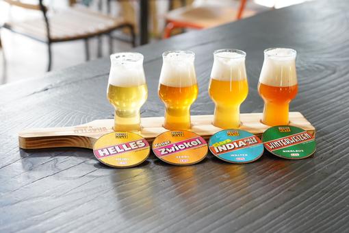 Das Foto zeigt vier Gläser unterschiedlicher Craft-Biersorten auf seinem hölzenen Tablett, davor färbige Bierdeckel mit der Bezeichnung der jeweiligen Biersorte