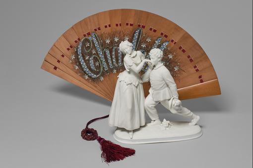 Small group of figures made of white porcelain with a woman and a man kneeling in front of her, behind them a reddish brown fan 