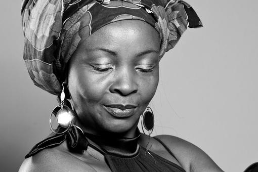 Black and white photo of the musician and artist Véro La Reine from Cameroon. She has her eyes closed and is wearing an elaborate fabric turban and eye-catching earrings