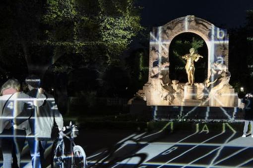 Das Foto zeigt das goldene Johann Strauss-Denkmal im Stadtpark in der Dunkelheit mit visuellen Effekten, im Vordergrund ein Paar, das am Denkmal vorbeigeht