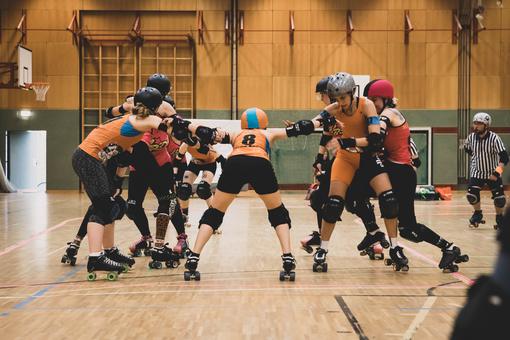 Photo of a Vienna Roller Derby game, women in orange jerseys, black shorts, with helmets and roller skates