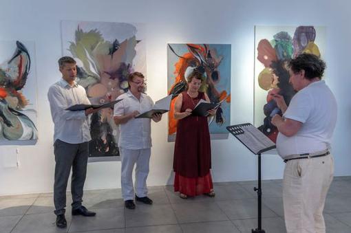 Three people singing and a conductor in front of them in a gallery with large pictures in the background