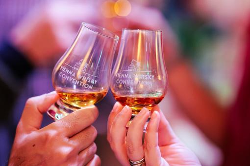 Foto von zwei mit Whiskey gefüllten Gläsern mit dem Logo des Vienna Whiskey Festivals, die so in Händen gehalten werden, um anzustoßen