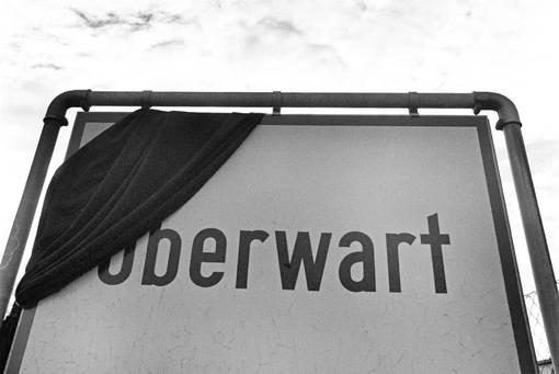 The photo shows the town sign of Oberwart with a mourning flag, in the background a sky covered with clouds