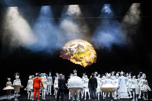 Szenefoto aus der Oper "Die Reise zum Mond", im Vordergrund zahlreiche Darsteller:innen, fast alle in weißen futuristischen Kostümen, die auf den Planeten Erde im Hintergrund blicken