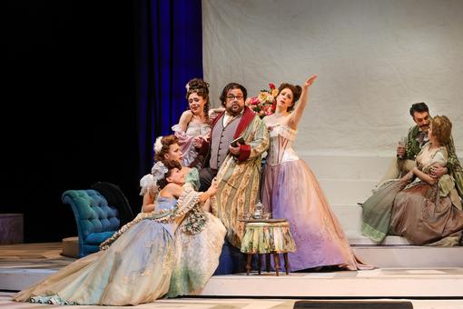 Szenefoto aus der Oper "La rondine", ein gut gekleideter Herr wird von vier Damen in wunderbaren Kleidern umringt