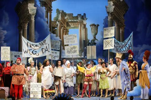 Scene photo from the operetta “Orpheus in the Underworld” with numerous performers in Greek mythology costumes and holding various posters as if at a demonstration