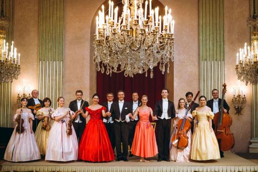Foto des Wiener Residenzorchesters, die Mitglieder in festlicher Abendrobe und mit Instrumenten, in einem historischen Saal mit großen Kronleuchtern