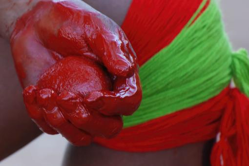 Der Fotoausschnitt zeigt die Vorbereitung zur Körperbemalung: eine in rote Farbe getauchte Hand, die einen roten Stein hält. Dahinter ein mit roten und hellgrünen Bändern umschlungenes Knie