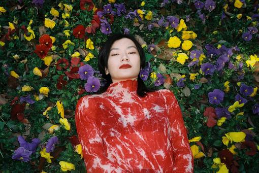 Zu sehen ist eine junge chinesische Frau in einem roten, weiß mamorierten Oberteil, die in einem Beet von lila, gelben und roten Stiefmütterchen liegt 