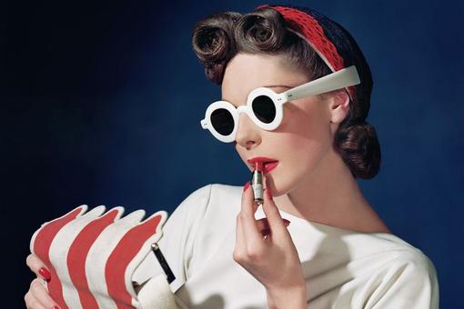 Photo of woman in sixty look with striking white sunglasses holding red lipstick to lips and holding white and red striped bag in other hand
