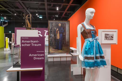 Exhibition view of a show about diversity with various exhibits such as photos, a dirndl dress, a historical painting showing a worker. In the foreground, a stand with the inscription American Dream