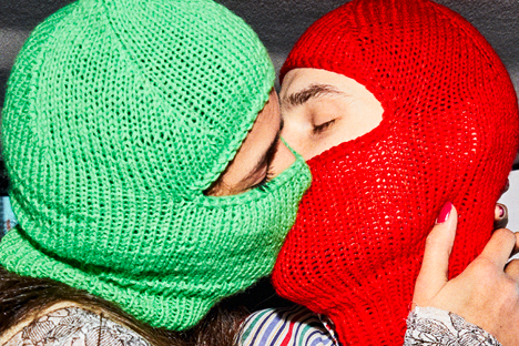 Das Foto zeigt zwei Personen, die sich küssen, beide tragen gestrickte Sturmhauben, eine in grellgün, die andere in rot, um ihre Gesichter zu verbergen